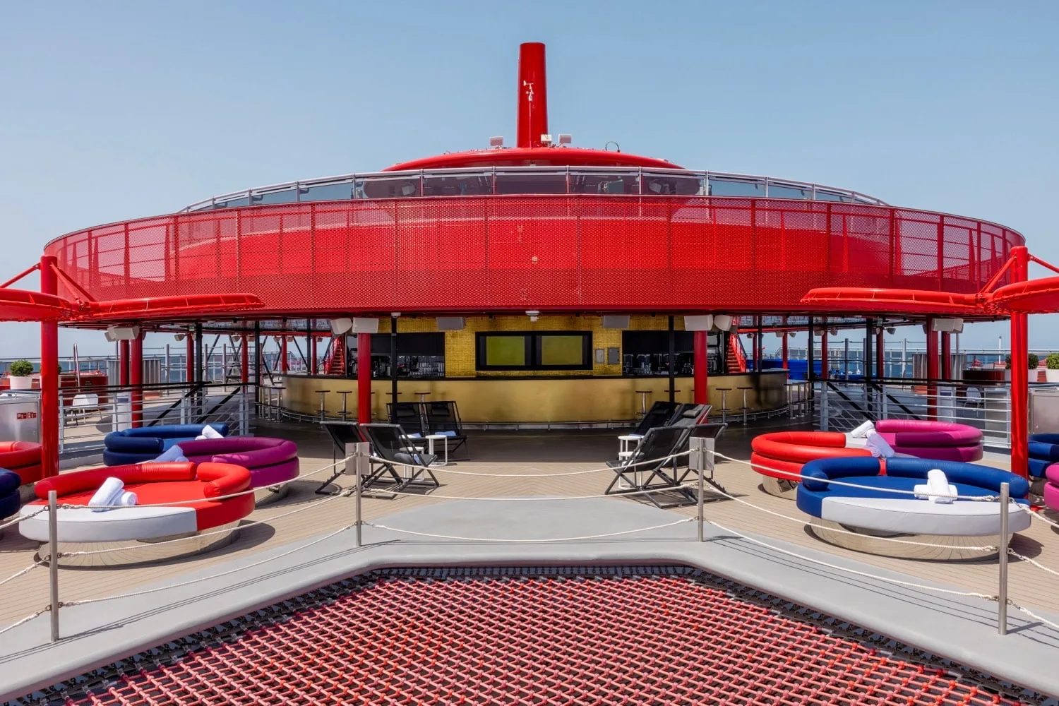 Outdoor lounge area on a ship with circular seating, tables, lounge chairs, and a red railing structure.