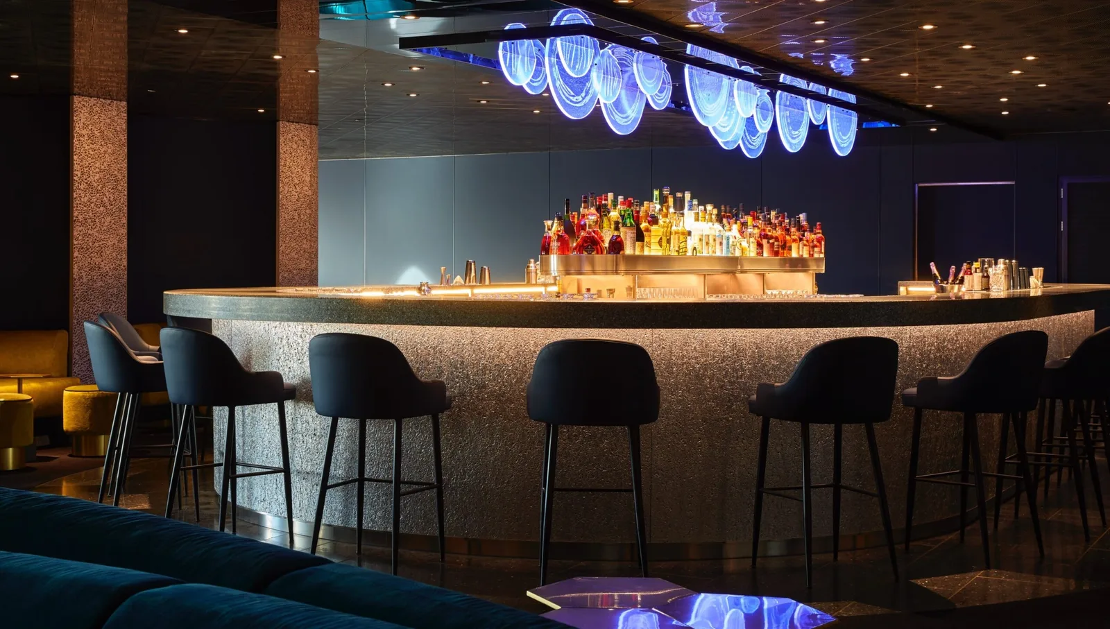 Modern onboard bar with illuminated blue circular ceiling lights, a variety of bottles displayed on the counter, and three bar stools.