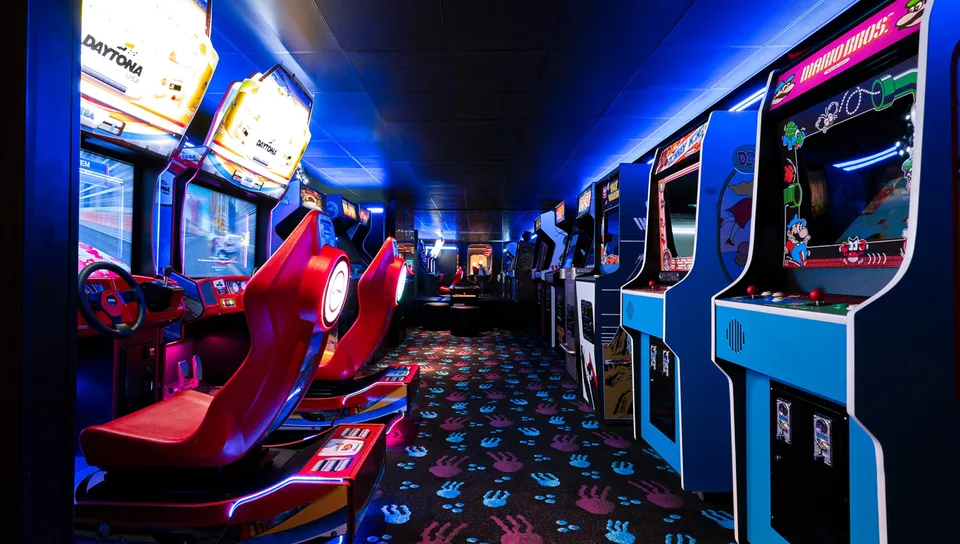 Arcade racing game machines with red seats and steering wheels are lined up in a dimly lit room with patterned carpet.