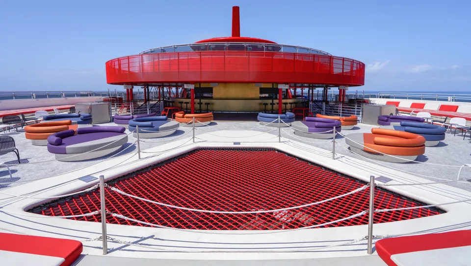 Outdoor deck area on a ship featuring circular seating and a red safety net, with a red circular upper deck structure in the background.