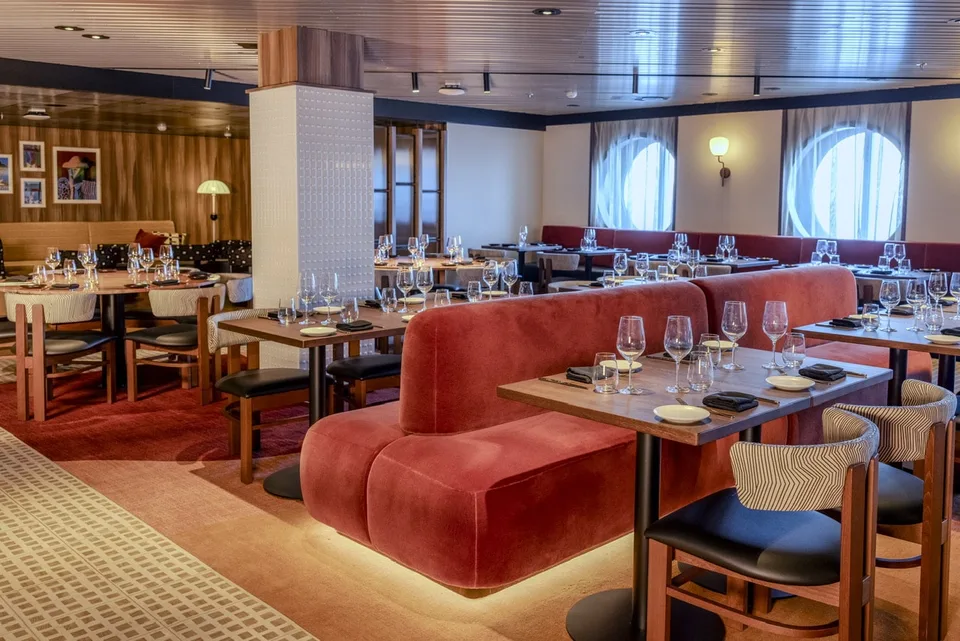 Interior view of a ship's dining area with red upholstered seating, wooden tables, chairs, and round porthole windows.