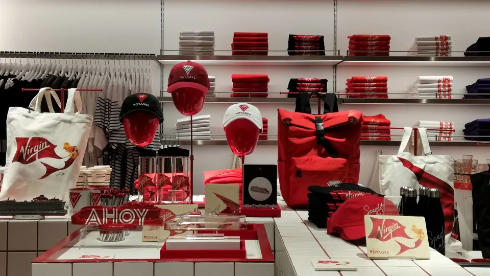 Display shelves with folded clothing, bags, a cap, and a Virgin Voyages promotional sign in a retail setting.