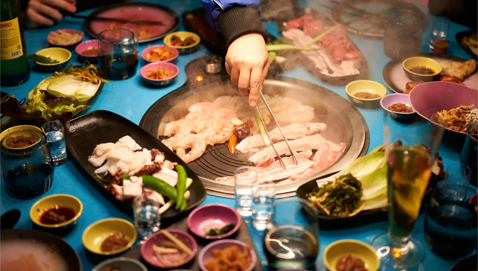 Overhead view of a person cooking shrimp and meat on a grill surrounded by various dishes and dipping sauces on a table.