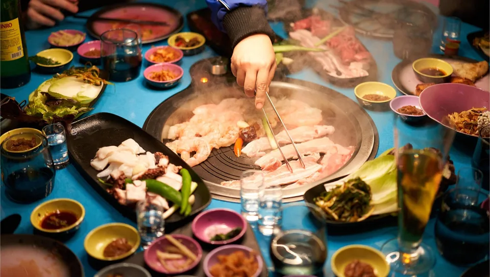 Overhead view of a person cooking shrimp and meat on a grill surrounded by various dishes and dipping sauces on a table.