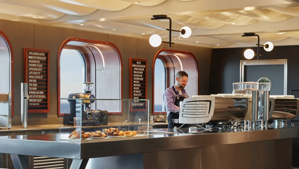 Interior view of a café or bakery area on a ship, featuring a glass display case with pastries and menu boards on the wall.
