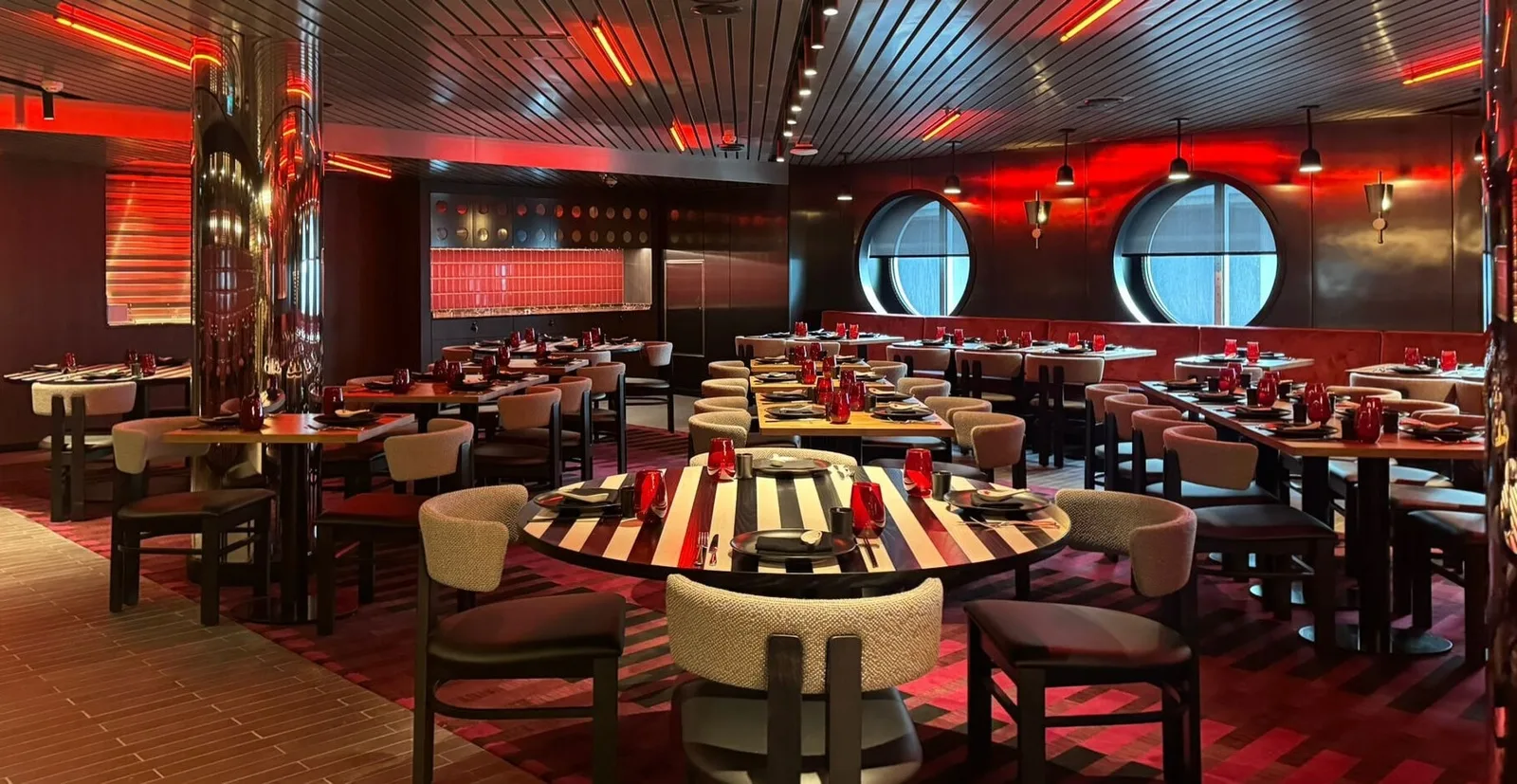 Interior of a stylish onboard restaurant with round tables, cushioned chairs, red lighting, and circular windows.