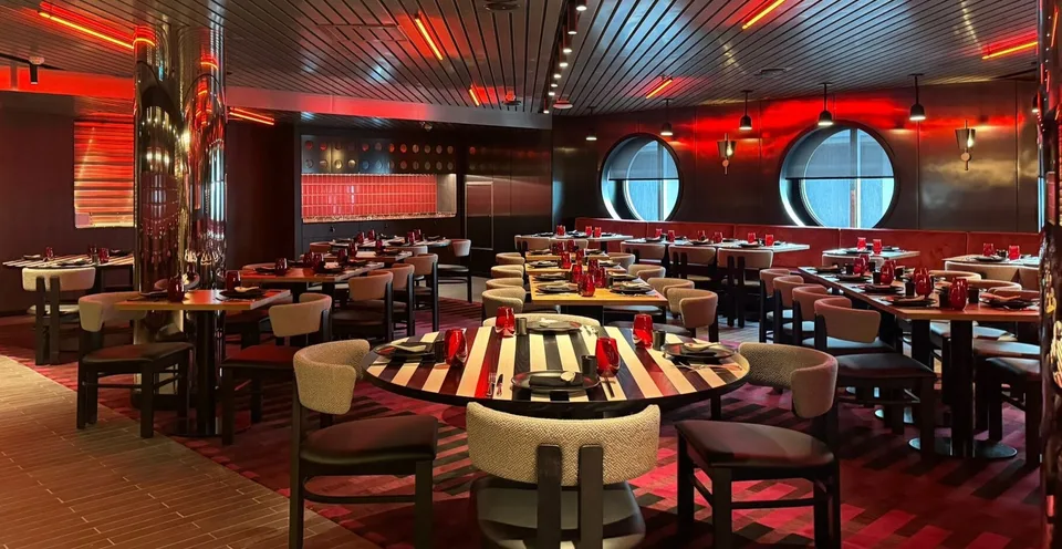 Interior of a stylish onboard restaurant with round tables, cushioned chairs, red lighting, and circular windows.