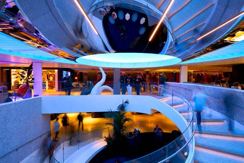 Interior view of a modern, multi-level space with curved staircases, glass railings, and a large circular chandelier or art installation on a cruise ship.
